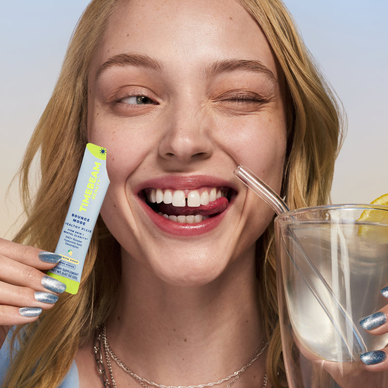 A smiling woman biting her tongue and winking while holding an open pouch of TIMEBEAM supplements and a glass of water with a straw.