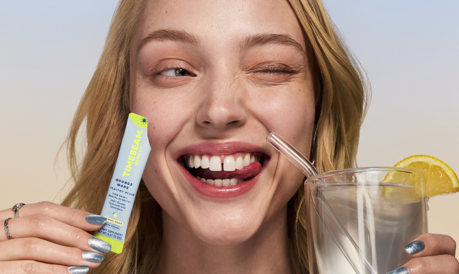A smiling woman holding a glass of water with a lemon slice and a TIMEBEAM Beauty Bounce Mode supplement pouch.