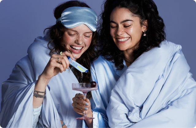 Two smiling women in blue blankets, one holding a drink while the other pours TIMEBEAM supplements into it.