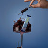Hand holding a skewer of cherries above a glass filled with cherries and a TIMEBEAM sleep supplement pouch.