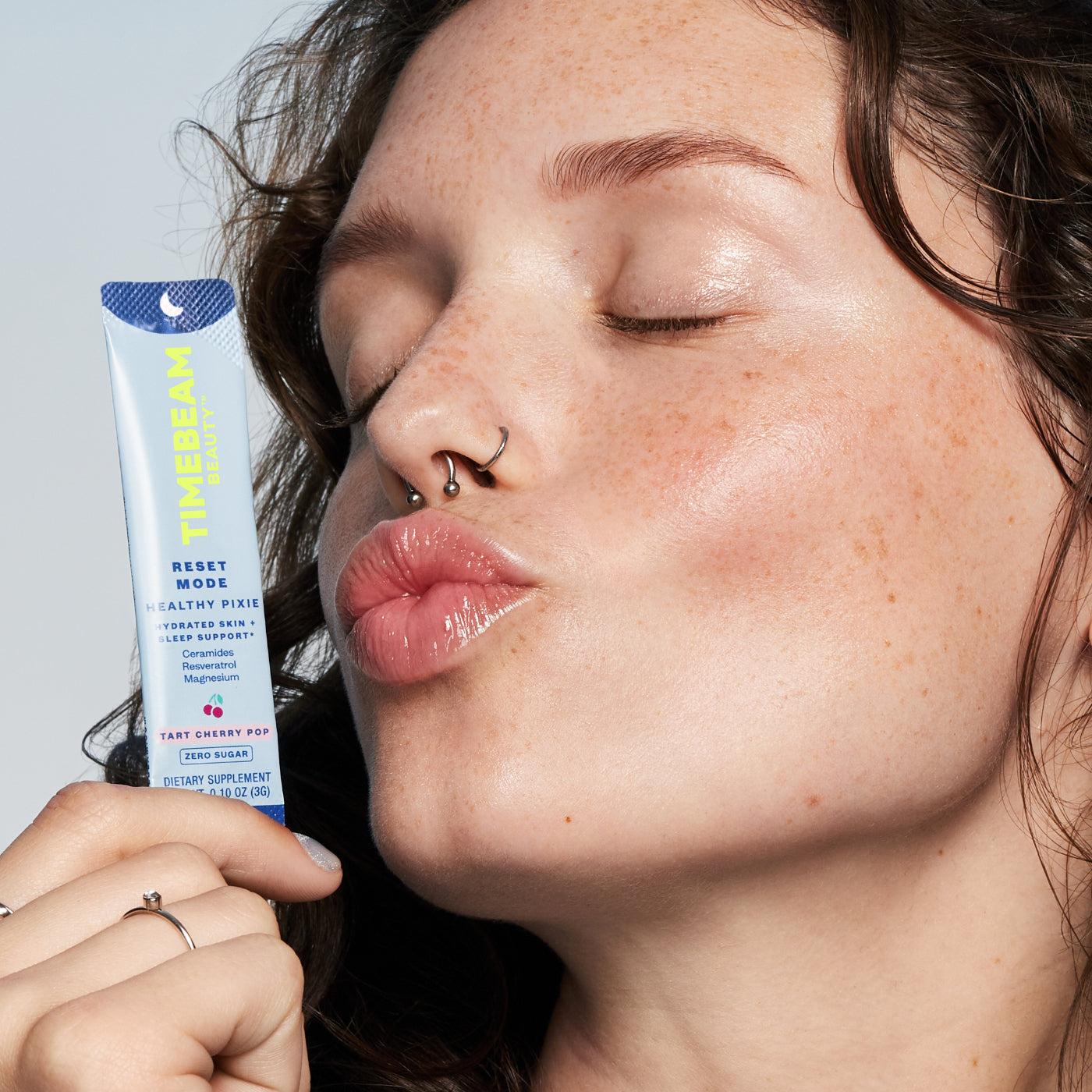 A woman holding a TIMEBEAM supplement pouch while making a kissing face with closed eyes.