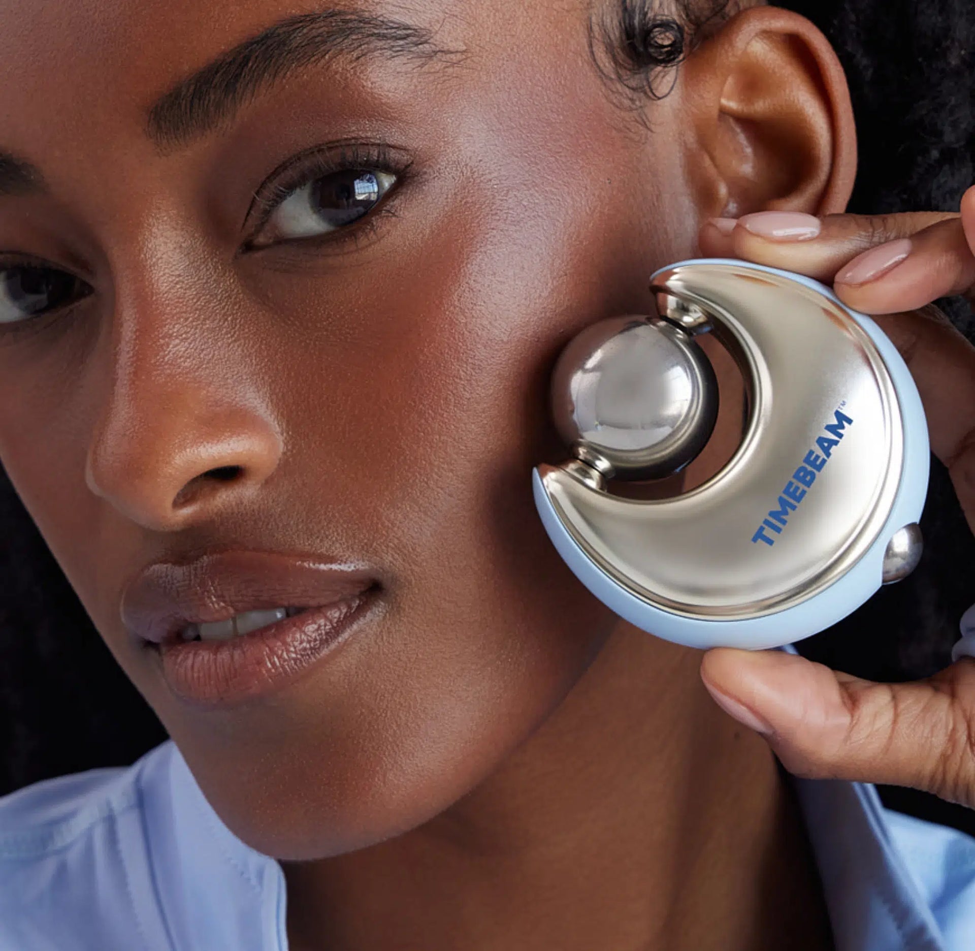 Close-up of a woman holding a silver and light blue TIMEBEAM facial device against her cheek.