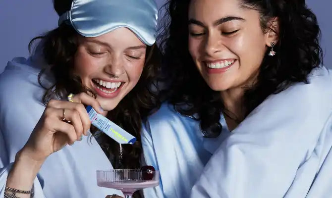 Two smiling women in blue blankets, one holding a drink while the other pours TIMEBEAM supplements into it.